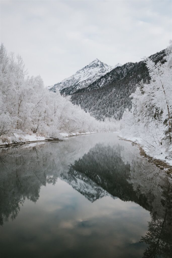 A snowy mountain landscape in Alaska showcases a river reflecting frosted trees and an overcast sky, inviting you to embark on a travel adventure.