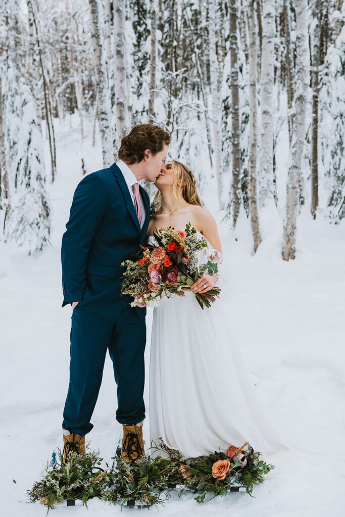 A couple in wedding attire kisses amidst a snowy forest during their winter elopement, both holding colorful flower bouquets.