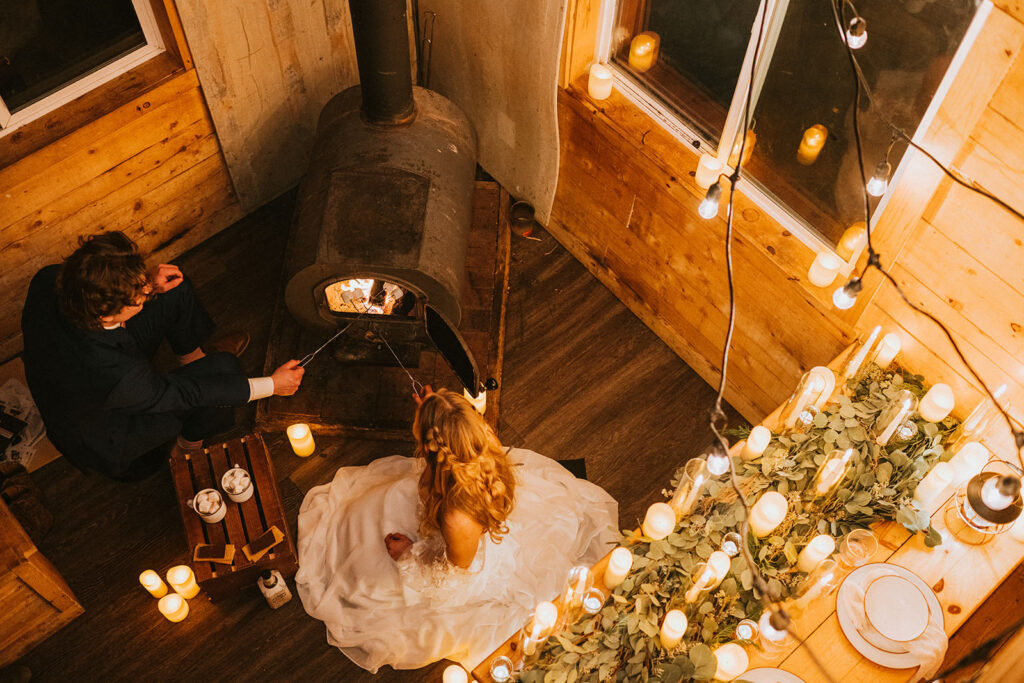 A bride and groom enjoy a cozy winter elopement by the fireplace in a wooden cabin, surrounded by candles and greenery on the table.