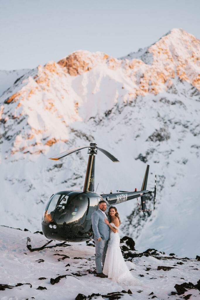 A couple in wedding attire stands on a snowy mountain, capturing the essence of a winter elopement with a helicopter poised in the background.