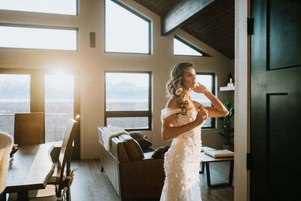 Bride in a white dress stands in a sunlit living room, adjusting her hair for the winter elopement. Large windows reveal a scenic mountain view outside.