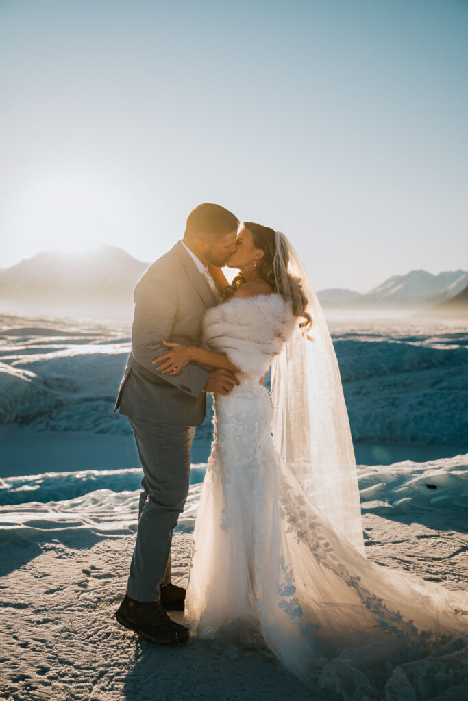 A couple in wedding attire shares a kiss on a snowy landscape during their winter elopement, with mountains in the background under a clear sky.