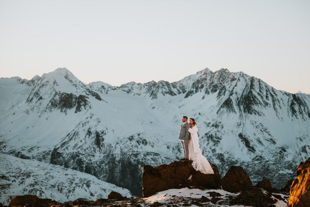 A couple in wedding attire stands on a rocky outcrop, embracing the spirit of winter elopement, as they overlook a snowy mountain range beneath a clear sky.