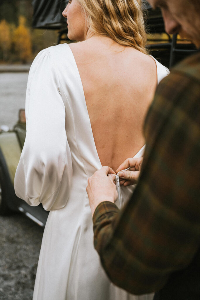 Person in a white dress has the back of the dress buttoned by another person outdoors.