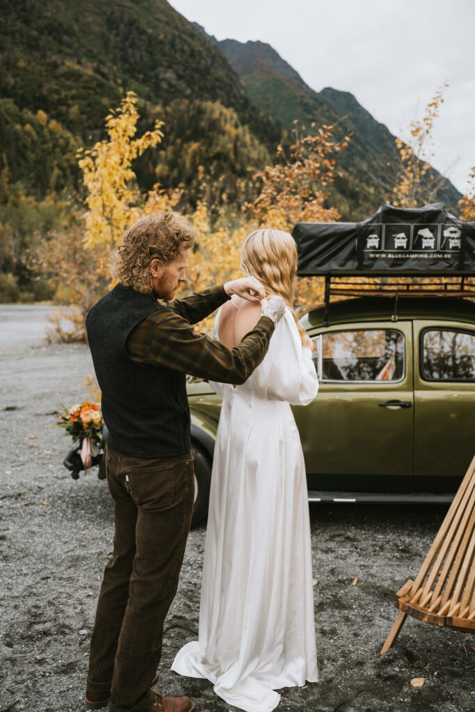 A person assists a woman in a white dress with her outfit near a green car in a scenic outdoor setting with mountains and autumn trees during their fall camping elopement.