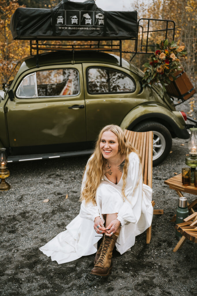 Woman in a white dress laces brown boots while sitting on a wooden chair beside a vintage green car with camping gear getting ready for her fall camping elopement.