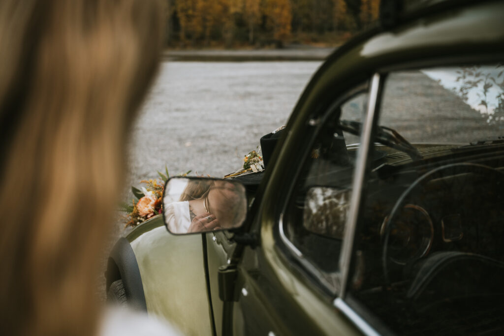 A person is visible through the side mirror of a vintage green car with flowers on its hood, set against an outdoor background.