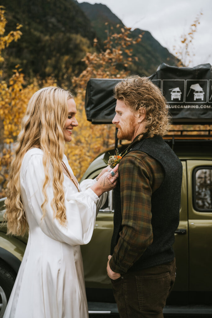 A woman in a white dress adjusts a boutonniere on a man's jacket. They stand in front of a vintage car with camping gear on top, surrounded by autumn foliage.