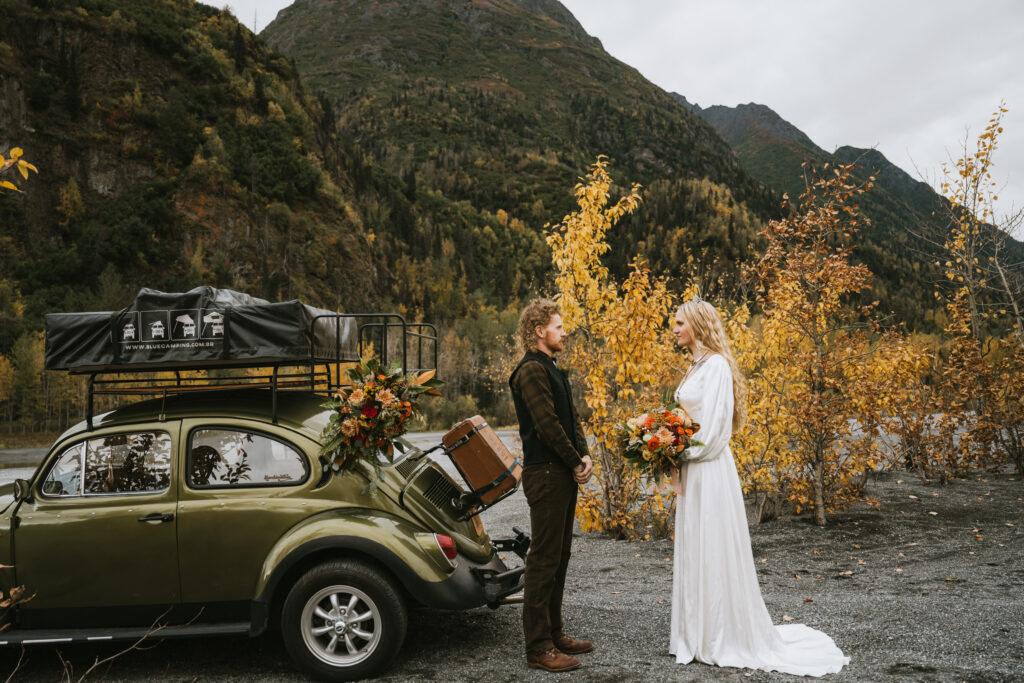 A couple stands by a vintage car with luggage on top, surrounded by autumn foliage and mountains. The woman wears a white dress, holding a bouquet.