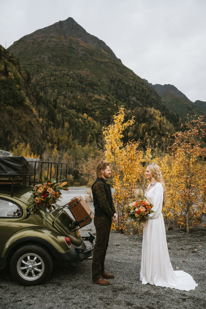 A couple stands facing each other in wedding attire, holding flowers, near a vintage car with luggage. Mountains and autumn trees are in the background.