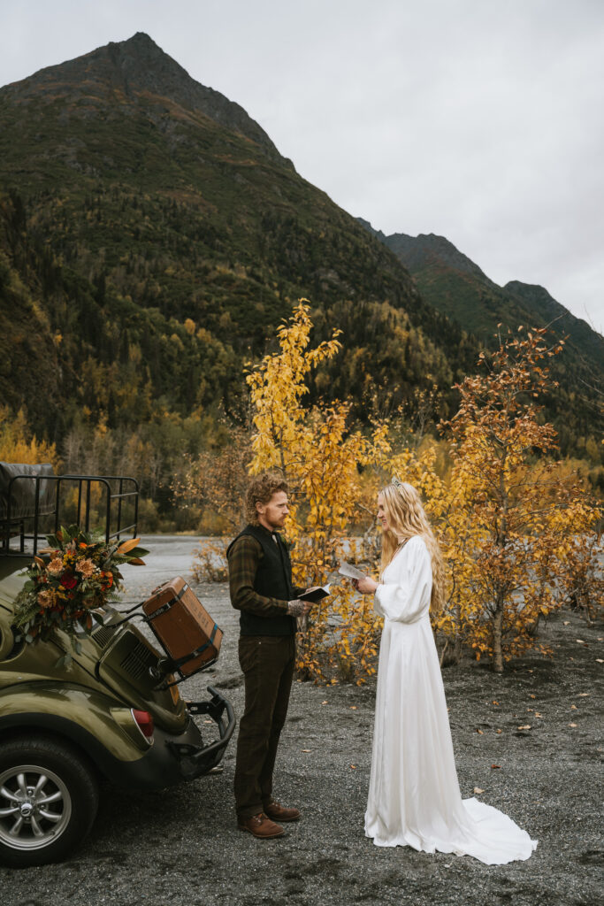 A couple stands facing each other, holding papers, in front of a vintage car with autumn foliage and a mountain in the background. The woman wears a long white dress.