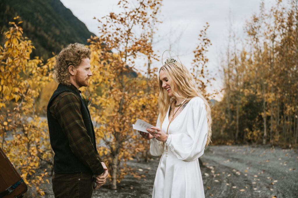 A couple stands outdoors during a ceremony. The woman reads from a card, while the man listens. They are surrounded by autumn trees and a mountain is visible in the background.