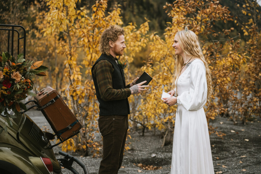 A couple stands outdoors exchanging vows, surrounded by autumn foliage. A vintage vehicle with luggage and flowers is beside them.