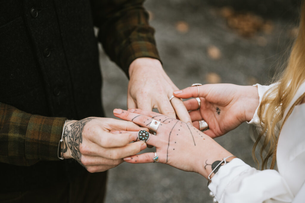 Close-up of two people exchanging rings, showing hands with tattoos and jewelry.