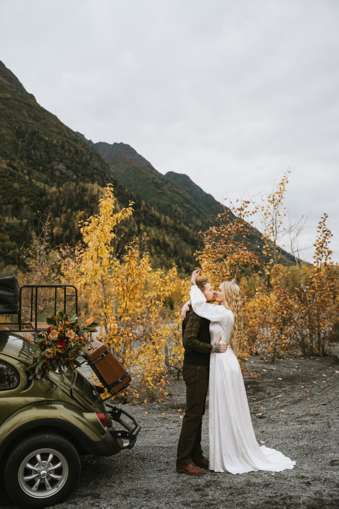 A couple embraces next to a green vintage car with a bouquet on the back. They're surrounded by autumn trees and mountains under a cloudy sky.