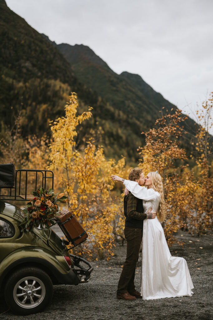 A couple embraces and kisses by an olive green car loaded with flowers and luggage, surrounded by autumn foliage and mountains.