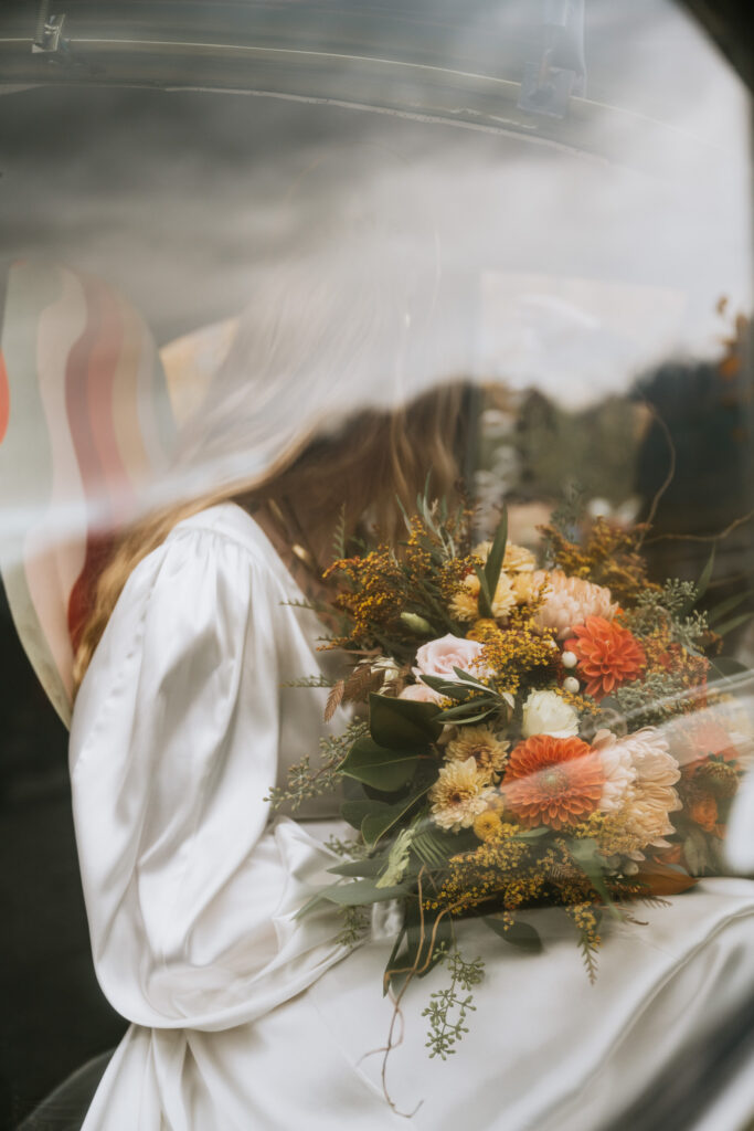 A bride in a white gown holds a large bouquet with various flowers, seen through a window.