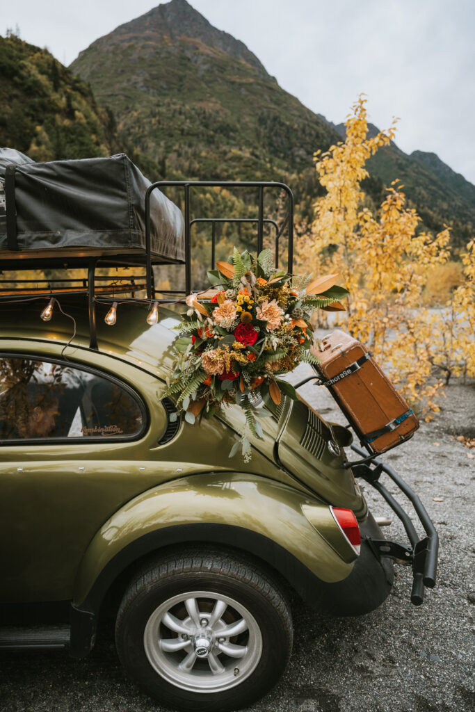 A green vintage car with luggage on the roof rack and a bouquet of flowers on the back is parked on a mountain road with autumn trees in the background.