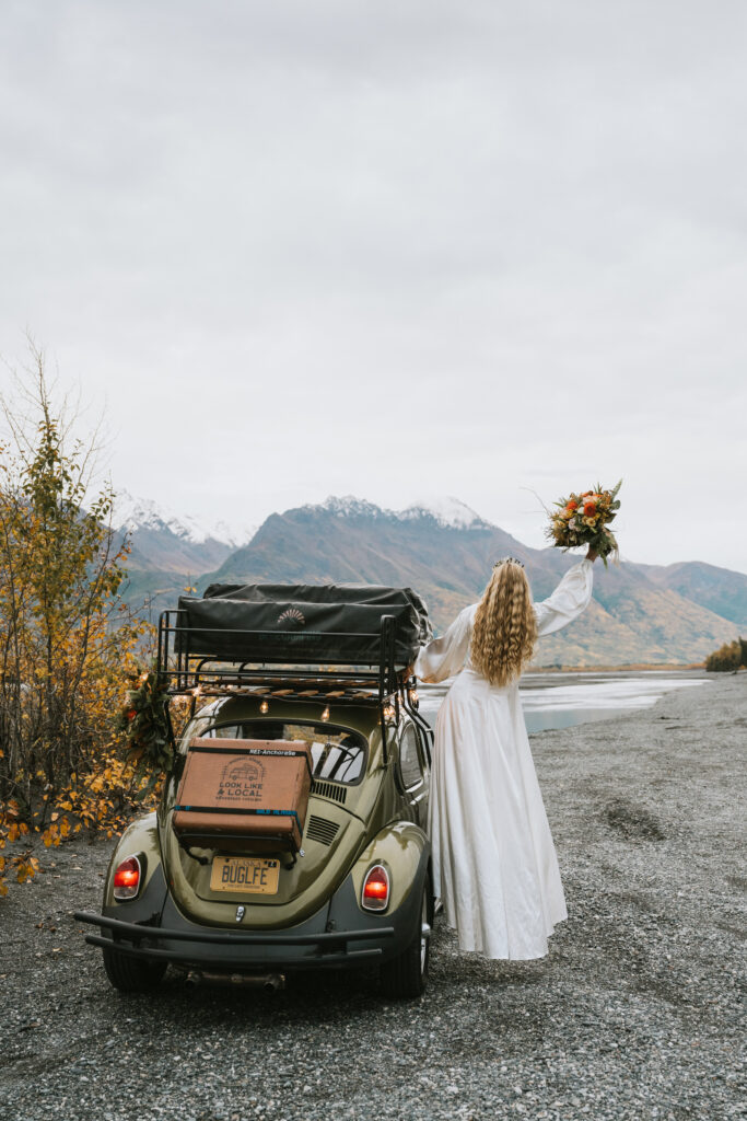 A person in a long white dress stands beside a vintage car with a roof rack, holding a bouquet while facing a scenic mountain and lake view.