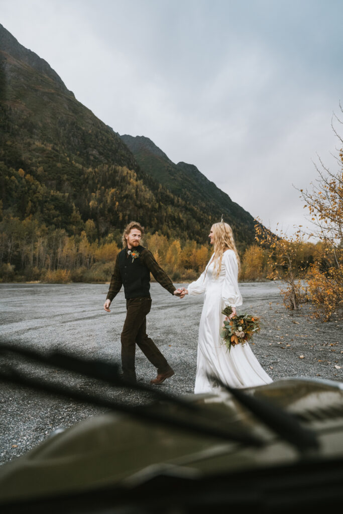 A couple in wedding attire holding hands and walking on a mountain road. The bride carries a bouquet, and autumn foliage is visible in the background.