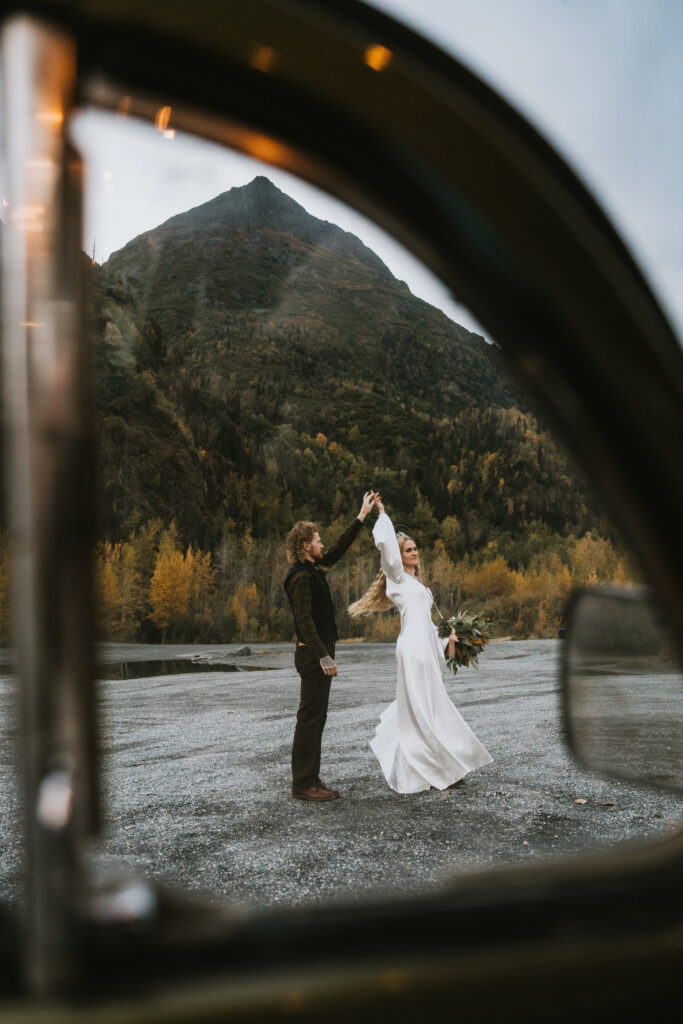 A couple dances outdoors near a mountain, framed by a car window. The woman is in a white dress holding a bouquet, and the man is in dark clothing.