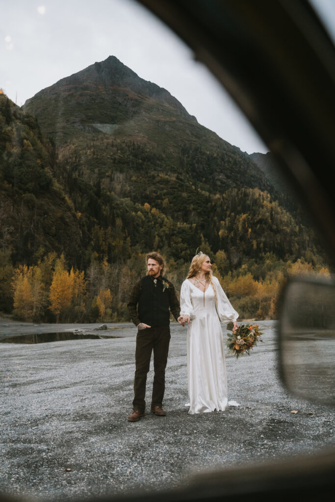 A couple stands on a mountain road, surrounded by autumn foliage. The woman holds a bouquet, both look into the distance. A car door frame partially visible in the foreground.