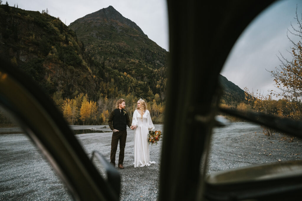 A couple stands holding hands in a mountainous landscape with autumn foliage, viewed through an open car door. The woman wears a white dress and holds a bouquet.