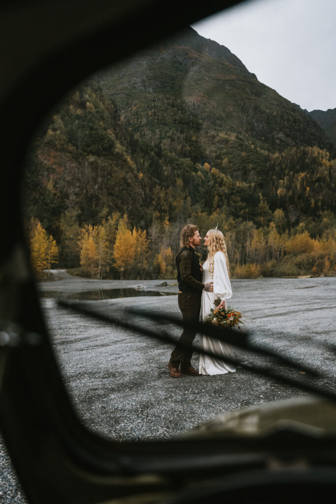 A couple in wedding attire stands closely on a gravel path surrounded by autumn trees, viewed through a vehicle window.