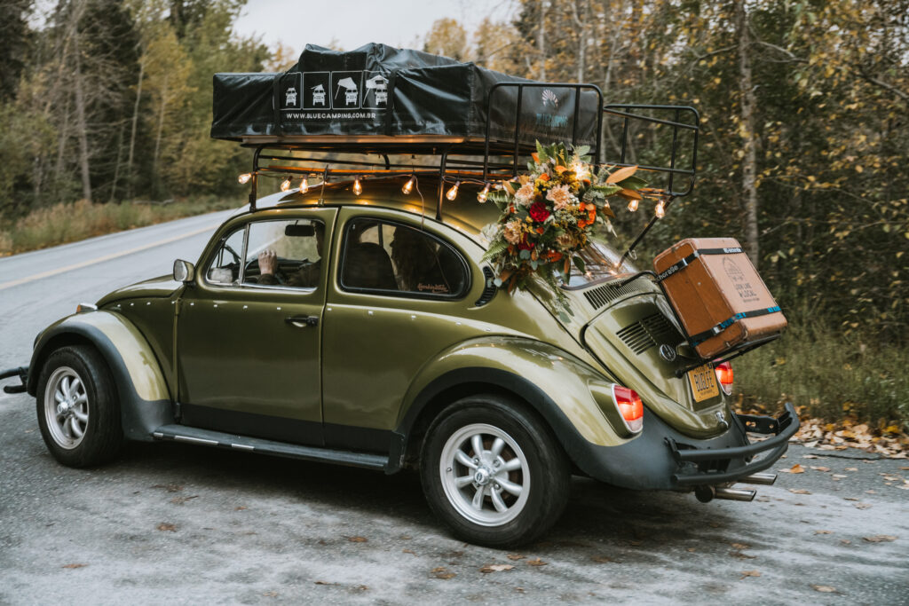 A vintage green car with a rooftop tent drives on a forested road. It is decorated with string lights and carries a bouquet and suitcase on the back.