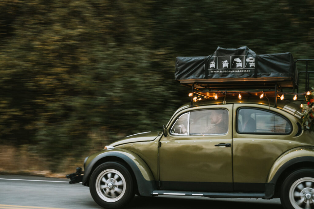 A green vintage Beetle with a roof rack drives on a road, surrounded by blurred greenery.