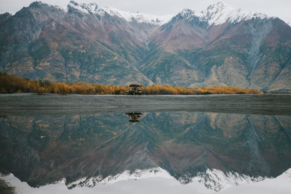 A vehicle on a flat landscape with snow-capped mountains and colorful autumn foliage reflected in a still body of water.