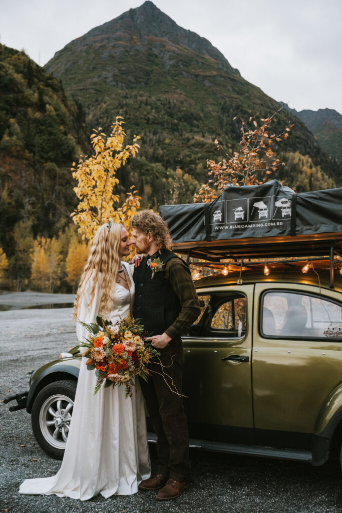 Bride and groom stand beside a vintage car with a rooftop tent. They are surrounded by autumn foliage, and a mountain is visible in the background. The bride holds a bouquet.