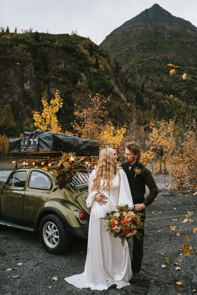 A couple in wedding attire stands by a flower-adorned vintage car in a scenic mountainous area with autumn foliage.