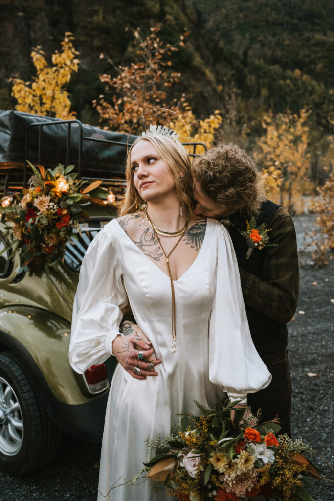 A couple in wedding attire stands by a decorated car. The woman holds a bouquet, and the man leans in close. Autumn foliage is visible in the background.