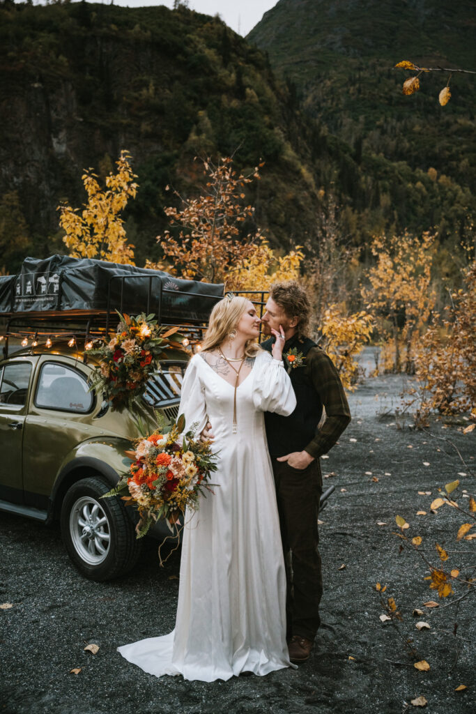 A couple embraces by a vintage car adorned with flowers. The woman is in a white dress, holding a bouquet, and the man is in a dark suit. Autumn foliage and a mountain form the background.