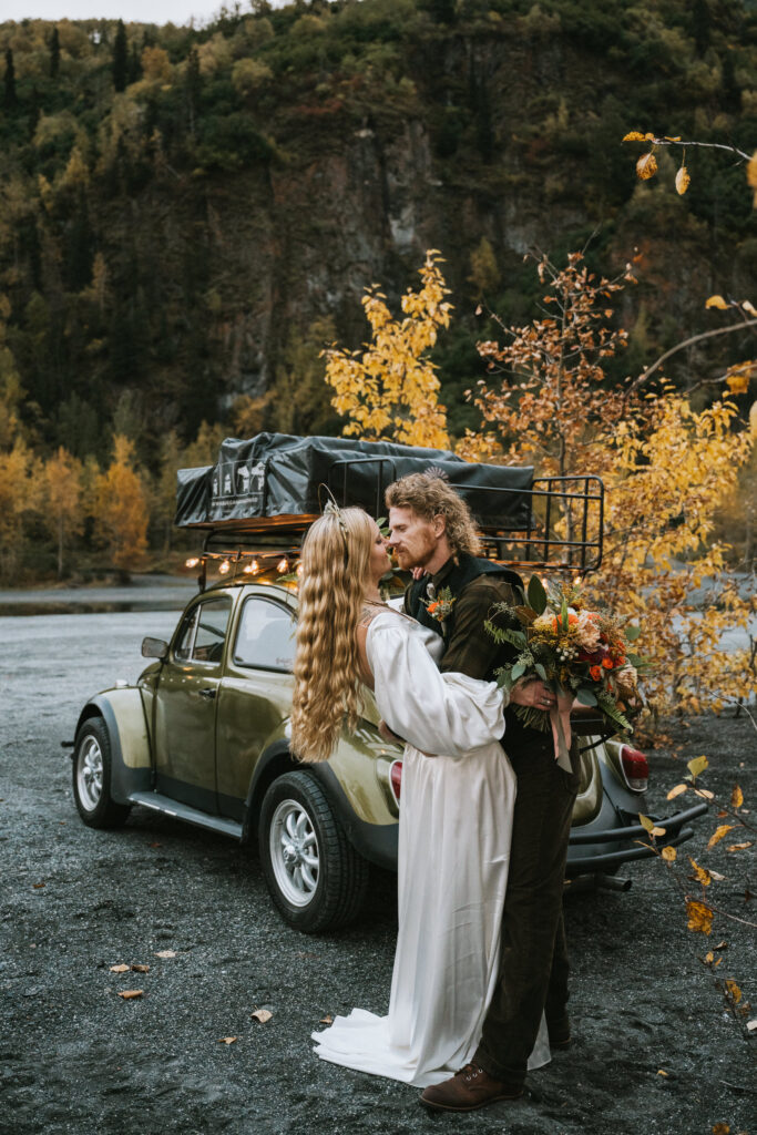 A couple in wedding attire kisses in front of a vintage car with a roof rack. They are surrounded by autumnal trees and a rocky background.