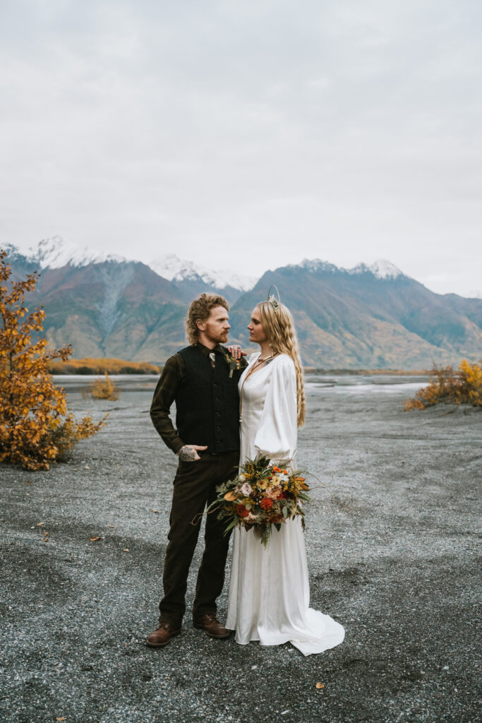 A couple stands on a gravel path with mountains in the background. The woman wears a long white dress and holds a bouquet, while the man wears a dark outfit.