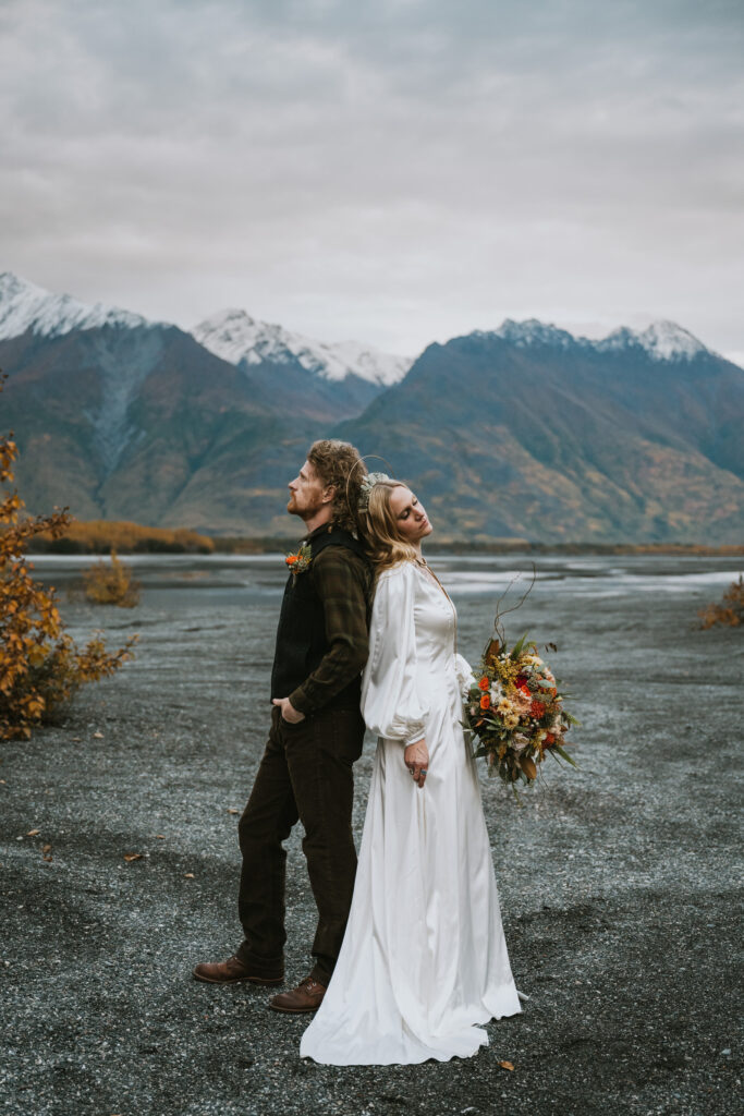 A couple in wedding attire stands back-to-back in a mountainous landscape with overcast skies. The bride holds a bouquet of flowers.