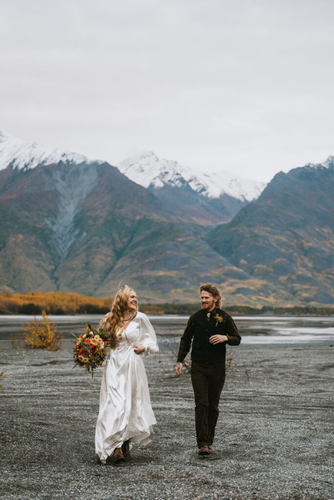 A couple walks outdoors, the woman in a white dress holding a bouquet, and the man in dark clothing. They are on a gravel path with mountains and trees in the background.