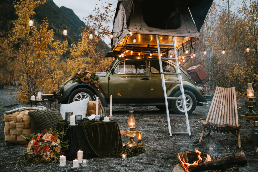 A vintage car with a rooftop tent is surrounded by outdoor furniture, candles, and a fire pit. String lights are hung across a wooded autumn setting for a fall camping elopement