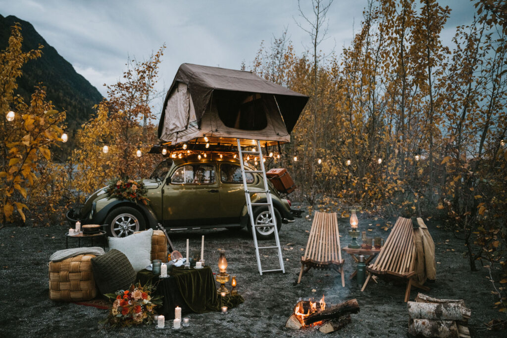 A car with a rooftop tent is set up for camping. Chairs and a fire pit are nearby, surrounded by autumn trees and string lights for a fall camping elopement.