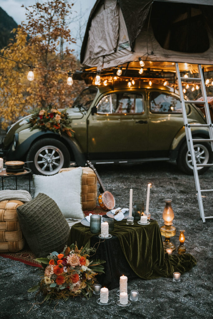 Vintage car with roof tent, surrounded by autumn foliage and lit string lights. Cozy setup with cushions, candles, and flowers in the foreground. Lanterns add a warm ambiance.