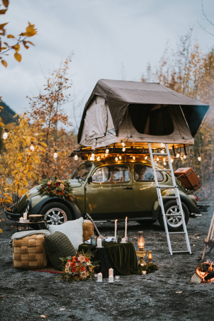 A vintage car with a rooftop tent, surrounded by autumn trees and outdoor camping decor, including string lights, cushions, and a small table with candles, fall camping elopement set up.