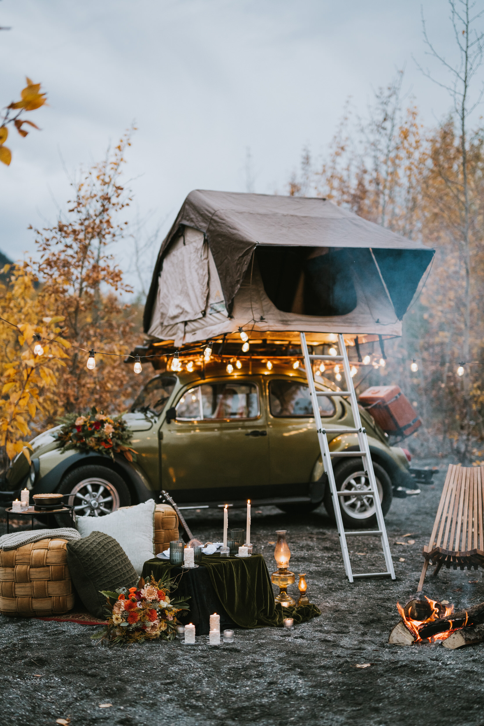 A vintage green car with a rooftop tent is set up for camping. It is surrounded by autumn trees, string lights, cushions, candles, and a small campfire.