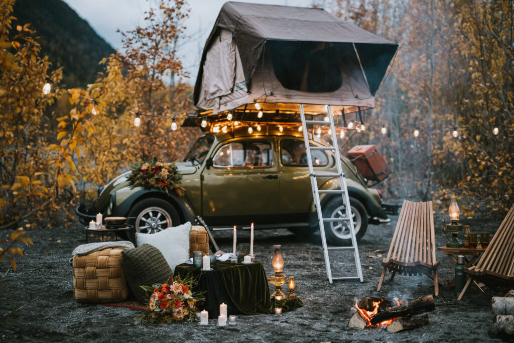 A vintage car with a rooftop tent is parked outdoors. Chairs, a fire pit, and string lights create a cozy campsite atmosphere amid autumn foliage.