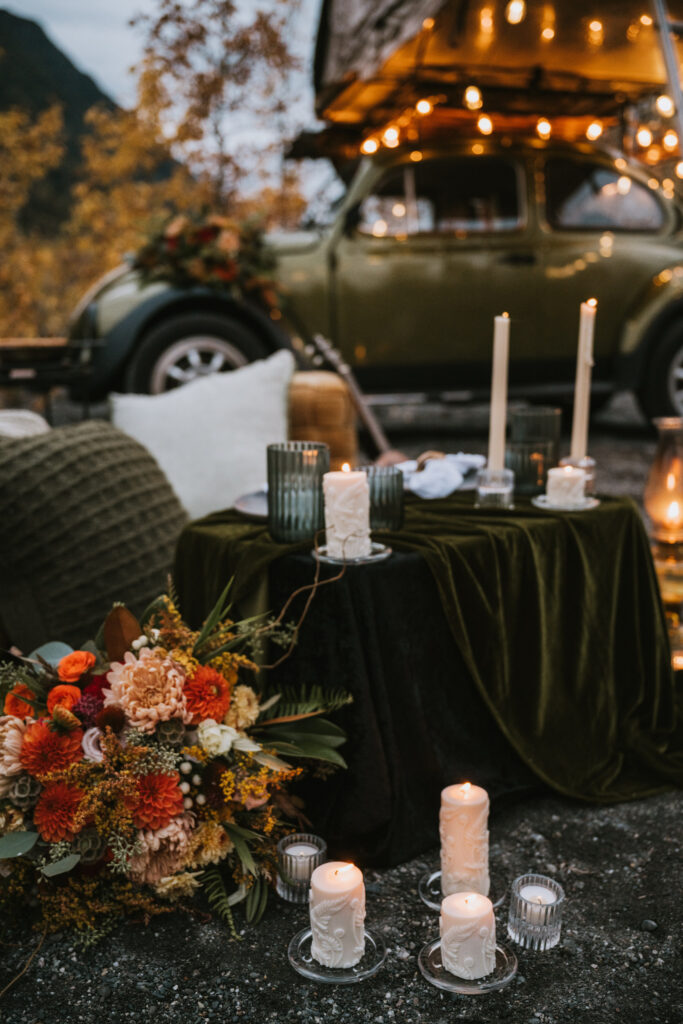 A cozy outdoor setting with a vintage car, illuminated by string lights. A table is adorned with candles, flowers, and velvet fabric, surrounded by pillows and greenery.