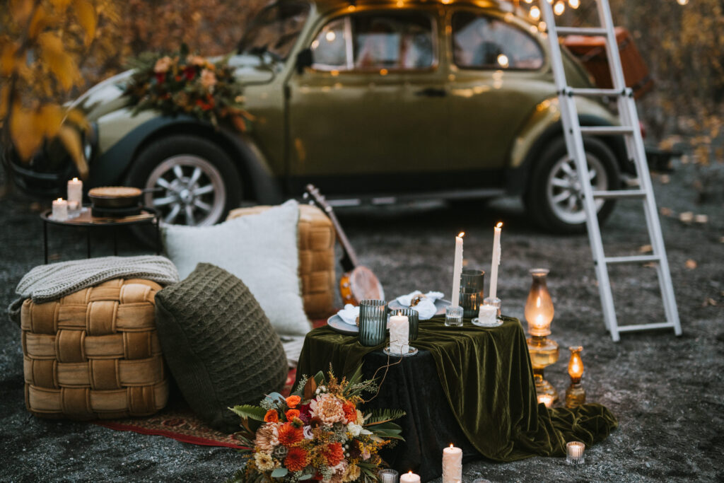 A vintage car is parked outdoors with a cozy seating area in front, featuring cushions, candles, a table with a green cloth, and a bouquet of flowers. A ladder leans against the car.