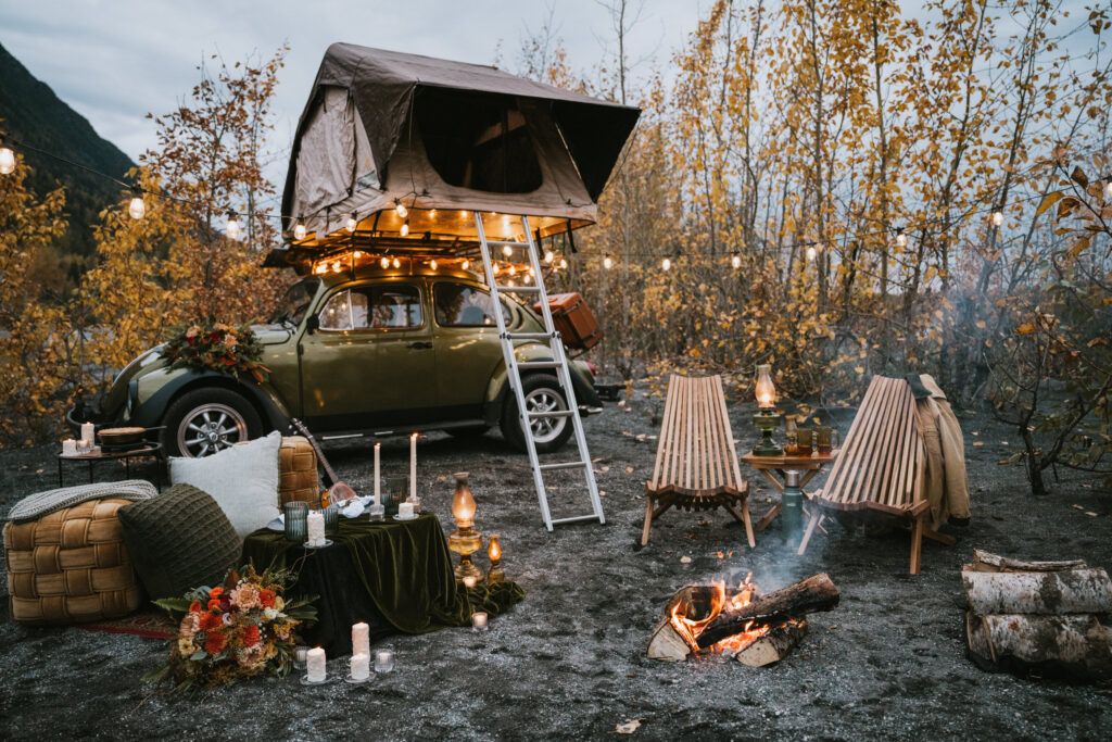 A vintage car with a rooftop tent is parked in a forest. A campfire is surrounded by two wooden chairs, baskets, and lanterns. String lights hang above in the trees.