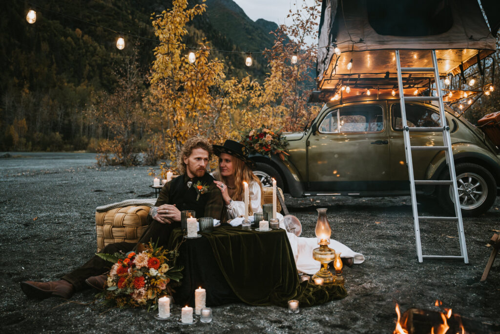 A couple sits on blankets with a small table, surrounded by candles and flowers. A vintage car with a rooftop tent is parked nearby, set against a backdrop of autumn trees and mountains.