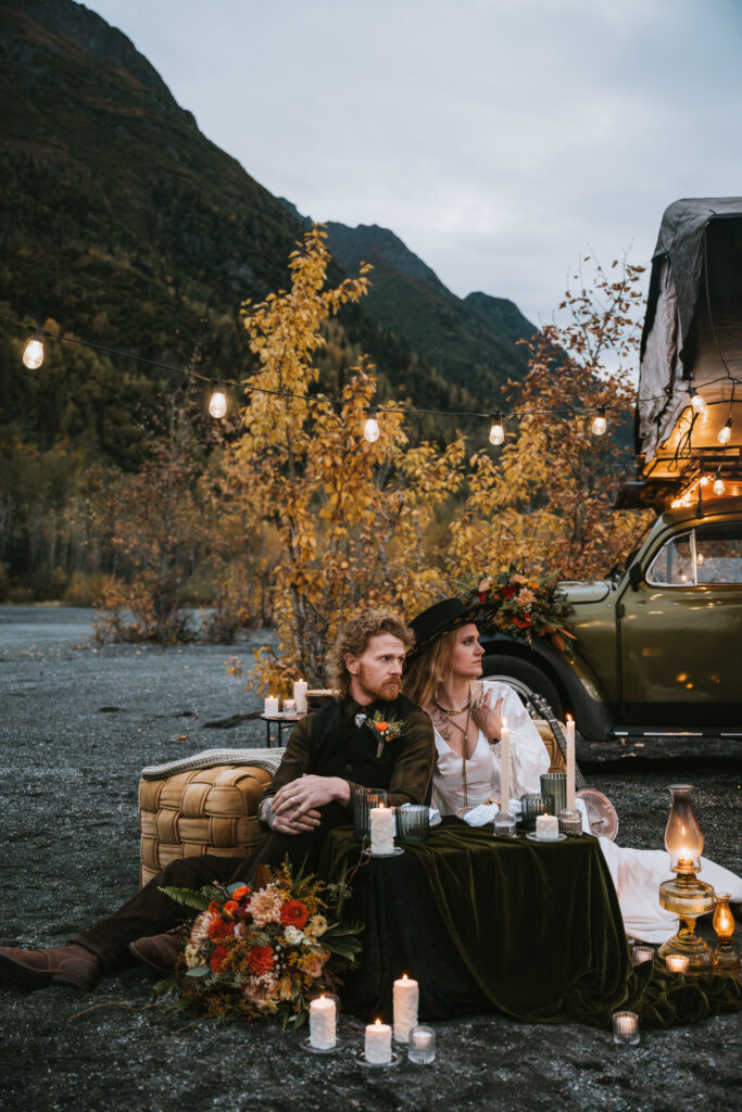 A couple sits outdoors on cushions near a table with candles and flowers, beside a vintage car. String lights hang above, with mountains and trees in the background.